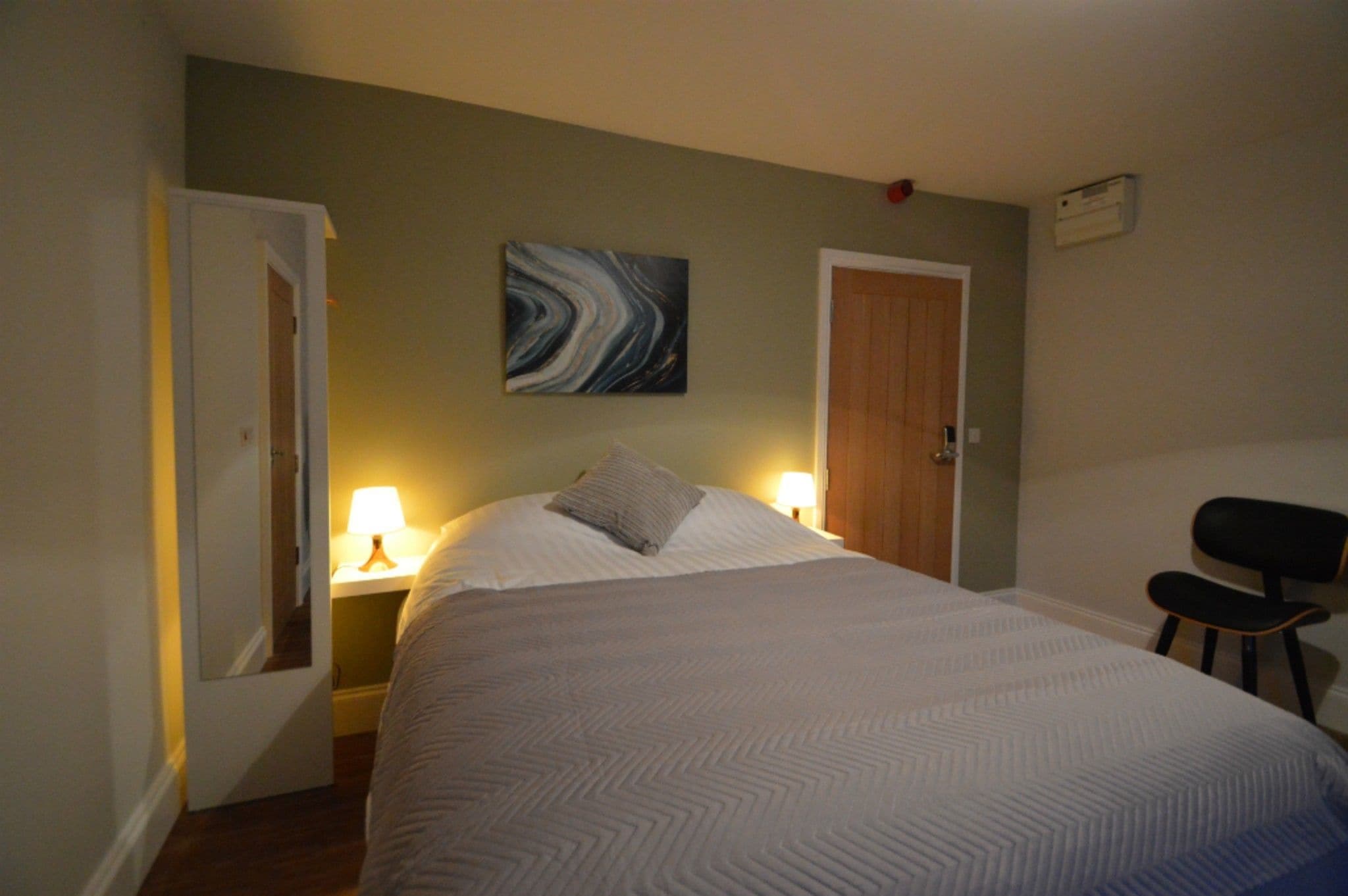 Un dormitorio con una cama, una mesita de noche con lámpara, una silla y una puerta.