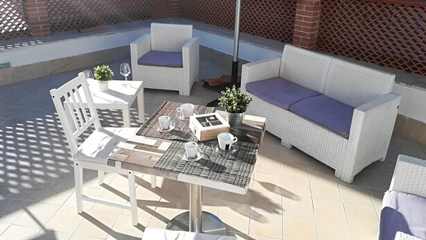 Een zonnig terras met een witte wickerbank, een witte tafel met glazen blad en een stoel.
