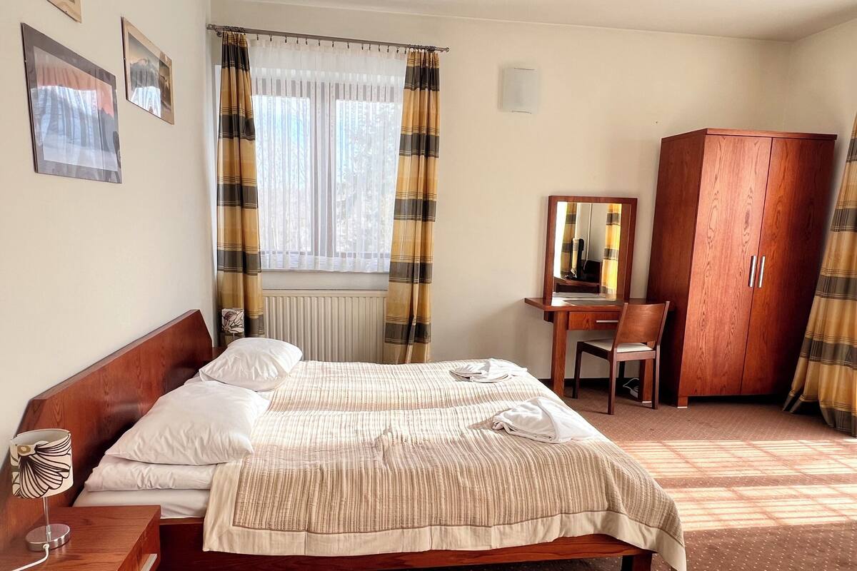 Habitación de hotel con una cama doble, una mesita de noche de madera, un escritorio con una silla, un armario y una ventana con cortinas.