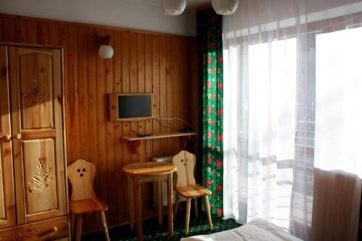 Una habitación con paneles de madera, una cama, un televisor montado en la pared, dos sillas de madera, una mesita y una ventana con cortinas translúcidas.