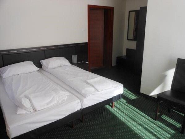 Cama doble con ropa blanca en habitación de hotel, con cabecera oscura, puerta de madera y un pequeño escritorio con silla.