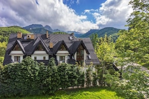 Una casa de estilo chalet con un techo pronunciado, múltiples chimeneas y fachada blanca. Está rodeada de exuberante vegetación y montañas al fondo.
