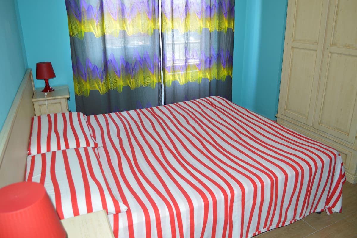 A bed with red and white striped bedding, a red lamp on a bedside table, and a window with purple and yellow curtains.