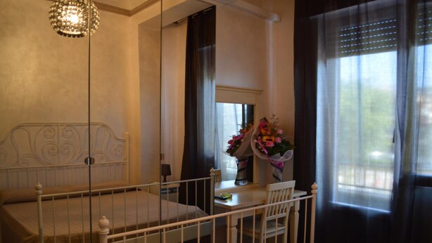 Ein Schlafzimmer mit Bett, Nachttisch, Stuhl, Fenster mit Vorhängen und einer Vase mit Blumen.