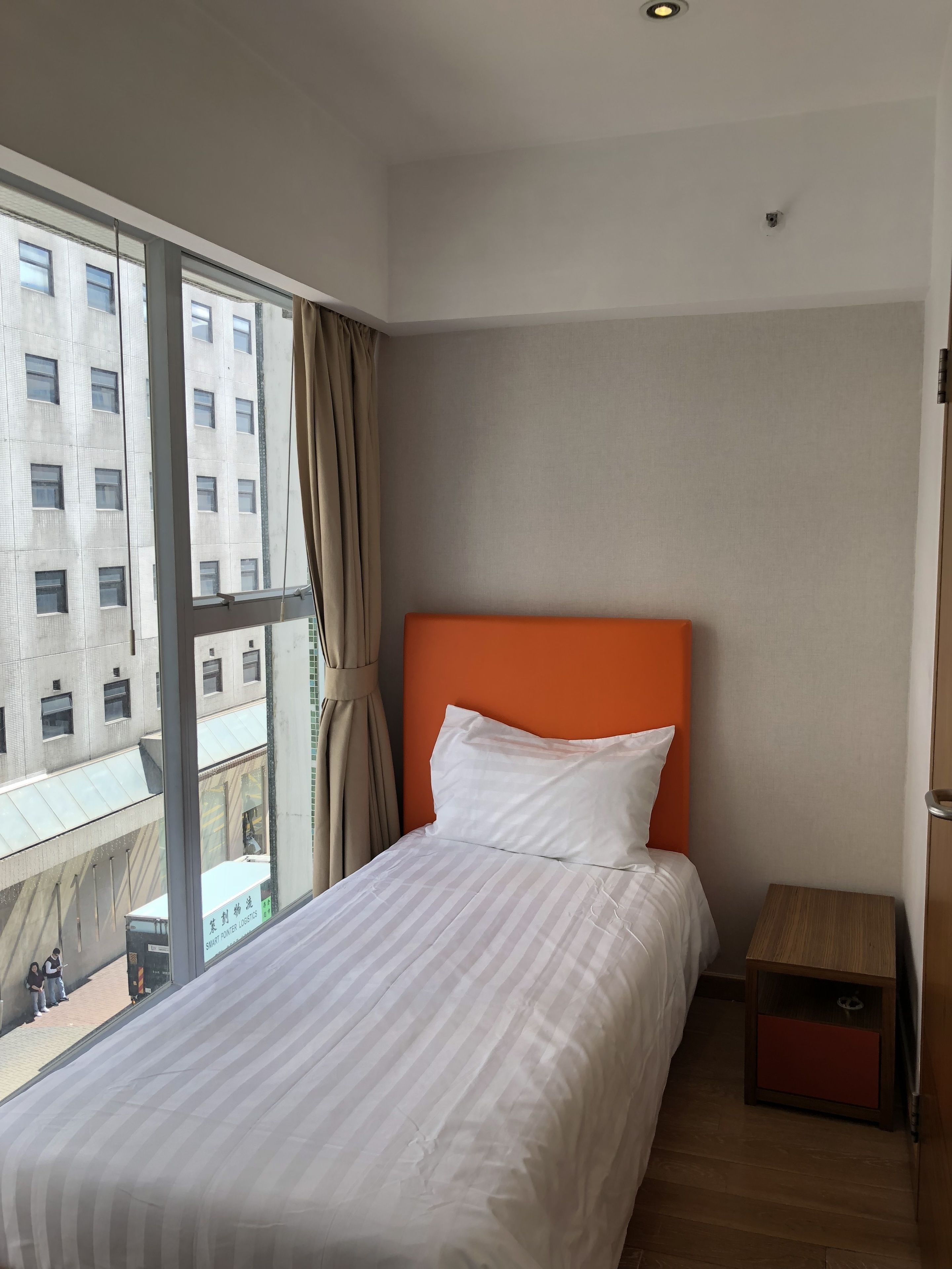 A hotel room with a large window, a bed with white bedding, an orange headboard, and a small wooden nightstand.