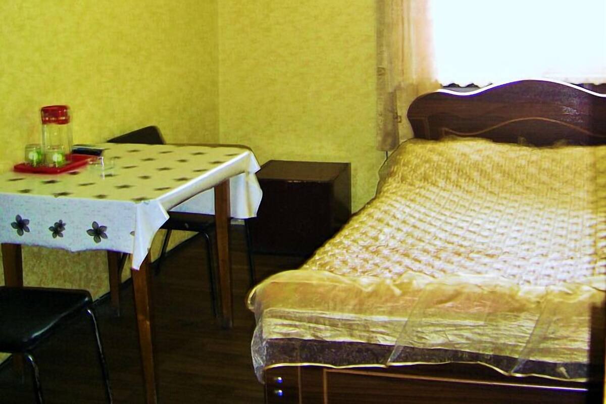 Une pièce comprenant un lit, une table avec des chaises, une petite table sur laquelle se trouve un récipient rouge, et une fenêtre avec des rideaux.