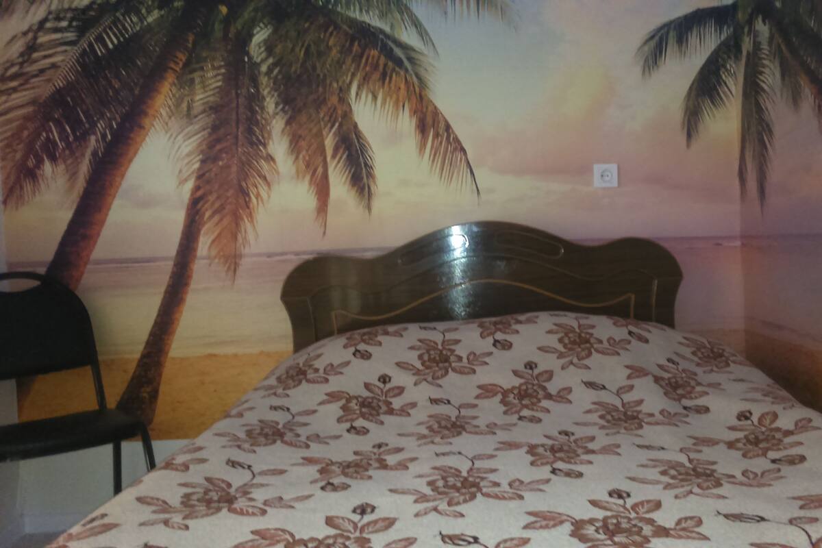 Un lit avec une couvre-lit à motifs floraux, une tête de lit en bois et une fresque murale représentant une scène de plage avec des palmiers.