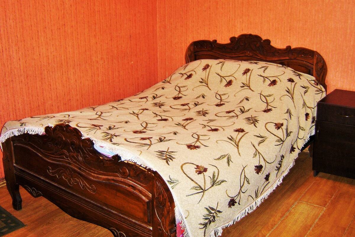 Un lit en bois, recouvert d’une couvre-lit à motifs floraux, dans une pièce aux murs orange et au plancher en bois.