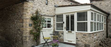 A stone house with a white door and windows, a small outdoor seating area with a table and chairs, and a potted plant with flowers.