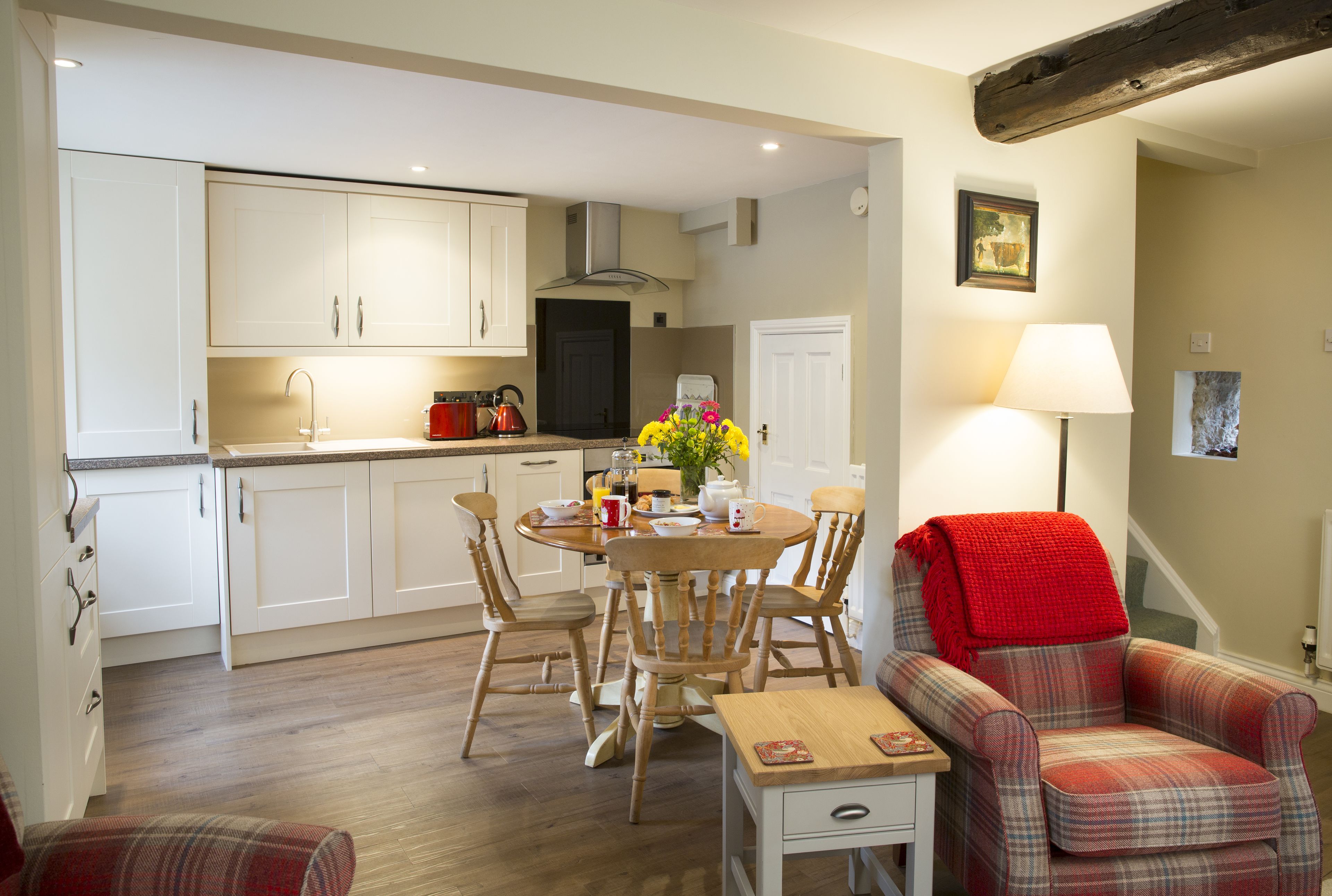 A compact kitchen with white cabinets, a wooden dining table with chairs, and a red plaid armchair.