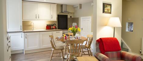 A compact kitchen with white cabinets, a wooden dining table with chairs, and a red plaid armchair.