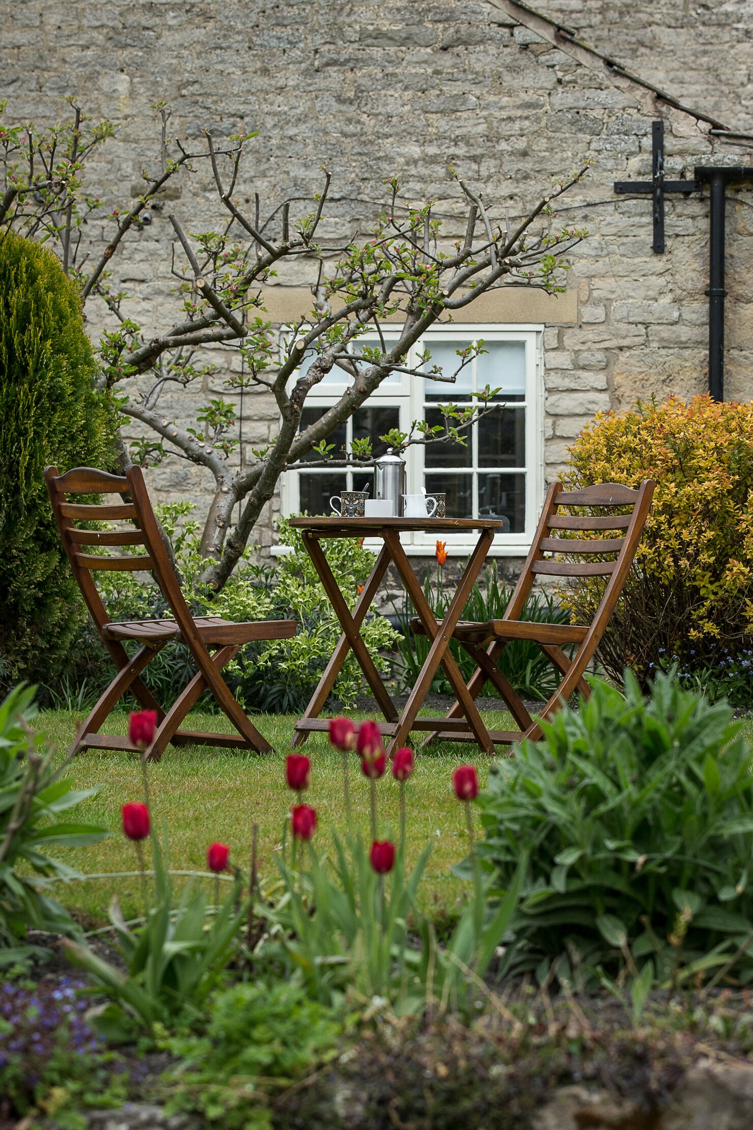 A garden with a stone wall, a wooden table, and two wooden chairs.