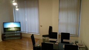 Un salon avec un téléviseur à écran plat posé sur un meuble, une table à manger en verre avec des chaises, et de grandes fenêtres équipées de stores.