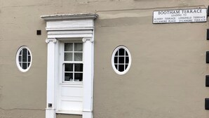 Une façade d’immeuble avec une porte blanche, deux fenêtres et un panneau indiquant les directions vers différentes terrasses.