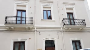 Un edificio de dos pisos con una puerta marrón, ventanas con persianas y un pequeño balcón con barandilla de hierro forjado negro.