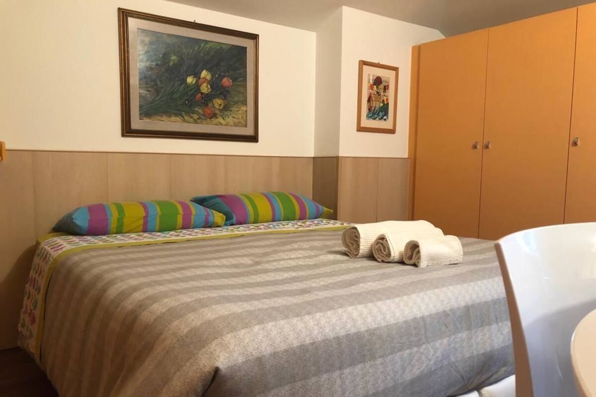 A hotel room with a bed, striped bedding, a colorful pillow, a painting on the wall, and a wardrobe.
