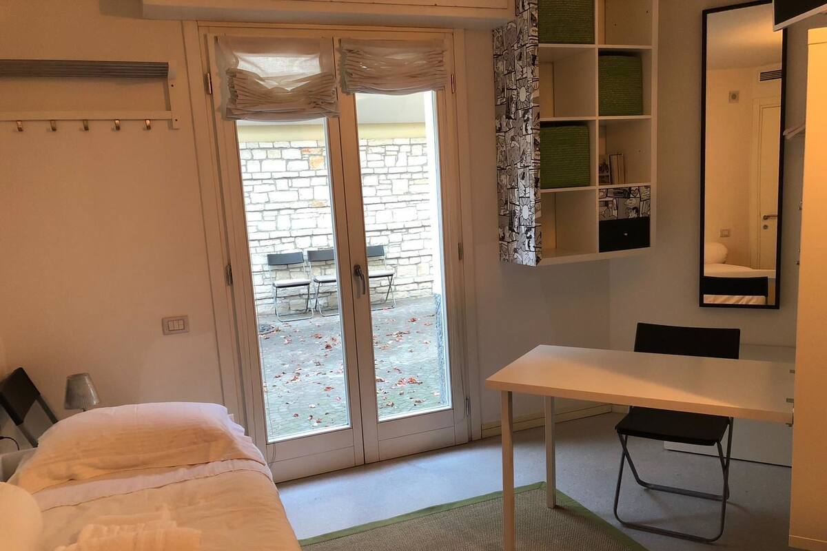 A hotel room with a bed, a desk, a chair, a mirror, and a view of an outdoor patio with chairs and a stone wall.
