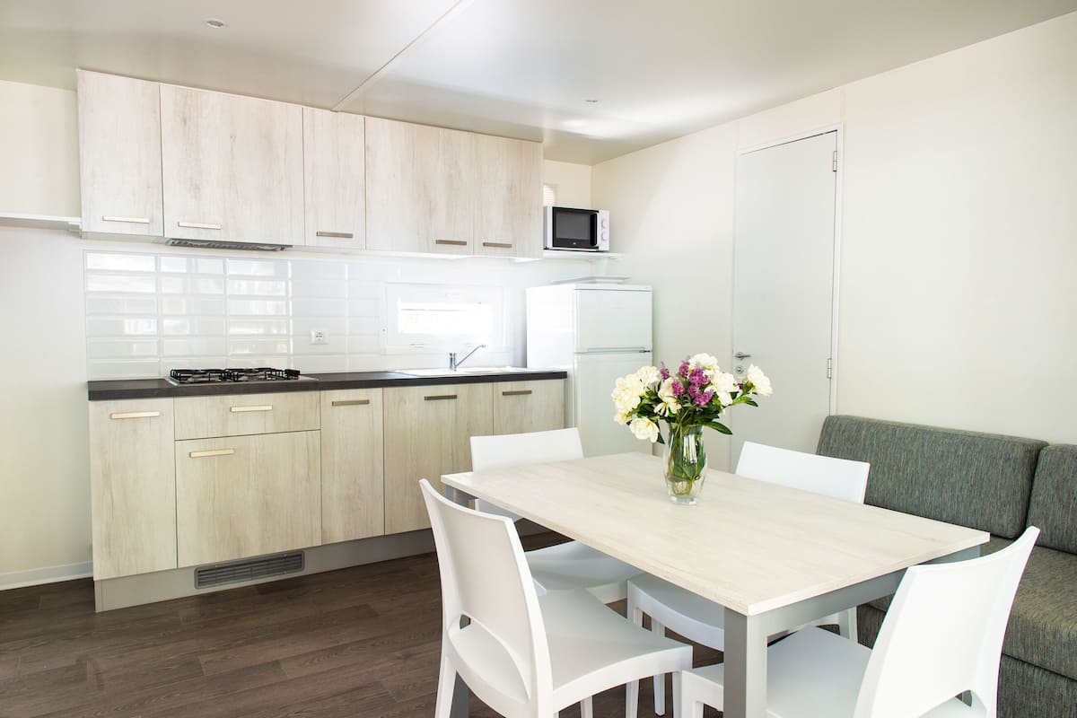 A modern kitchen with light wood cabinets, a white countertop, and a built-in microwave. There is a dining table with white chairs and a vase of flowers. A gray sofa is against the wall.