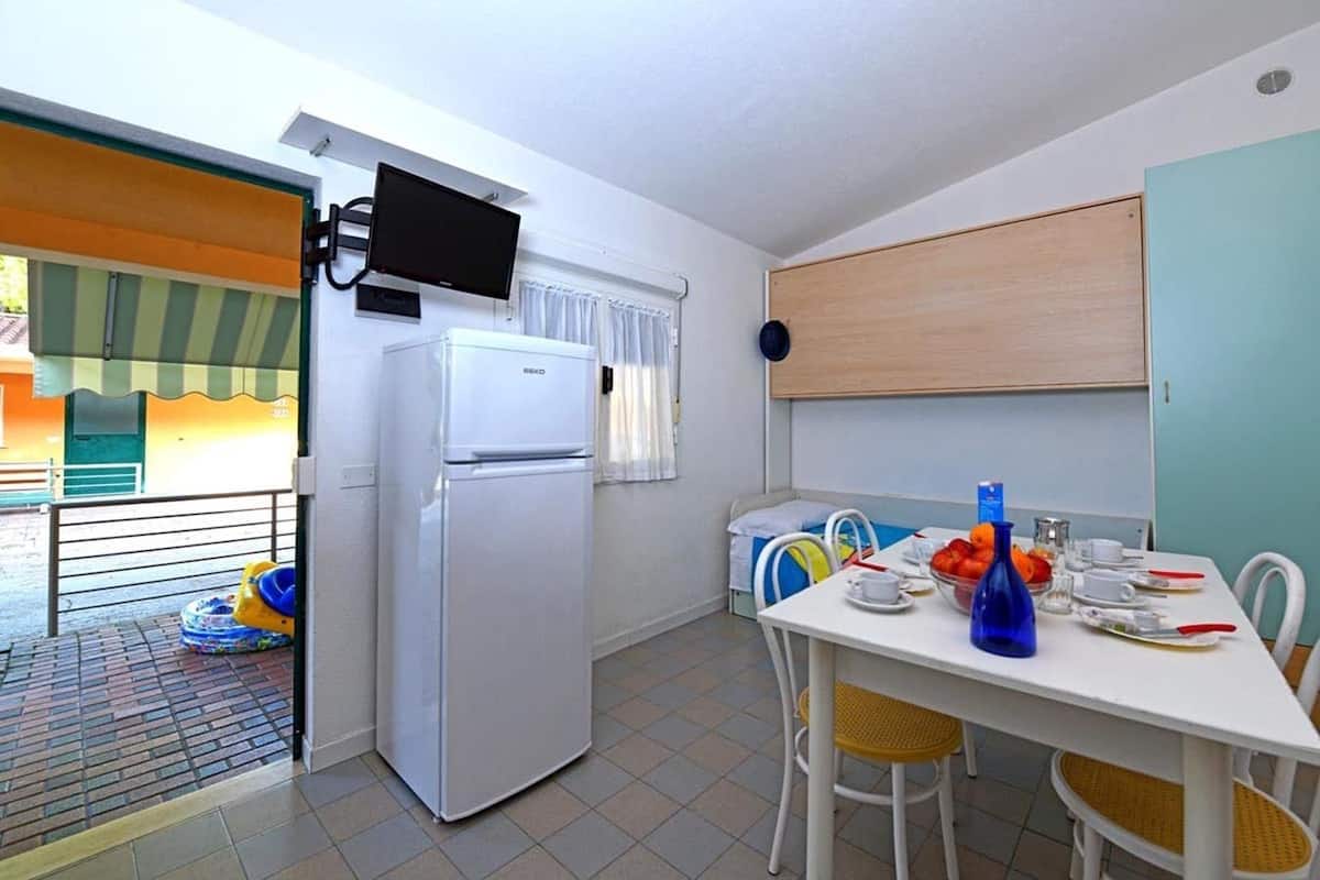 A compact kitchen with a refrigerator, a dining table set for two, and a small window with curtains.