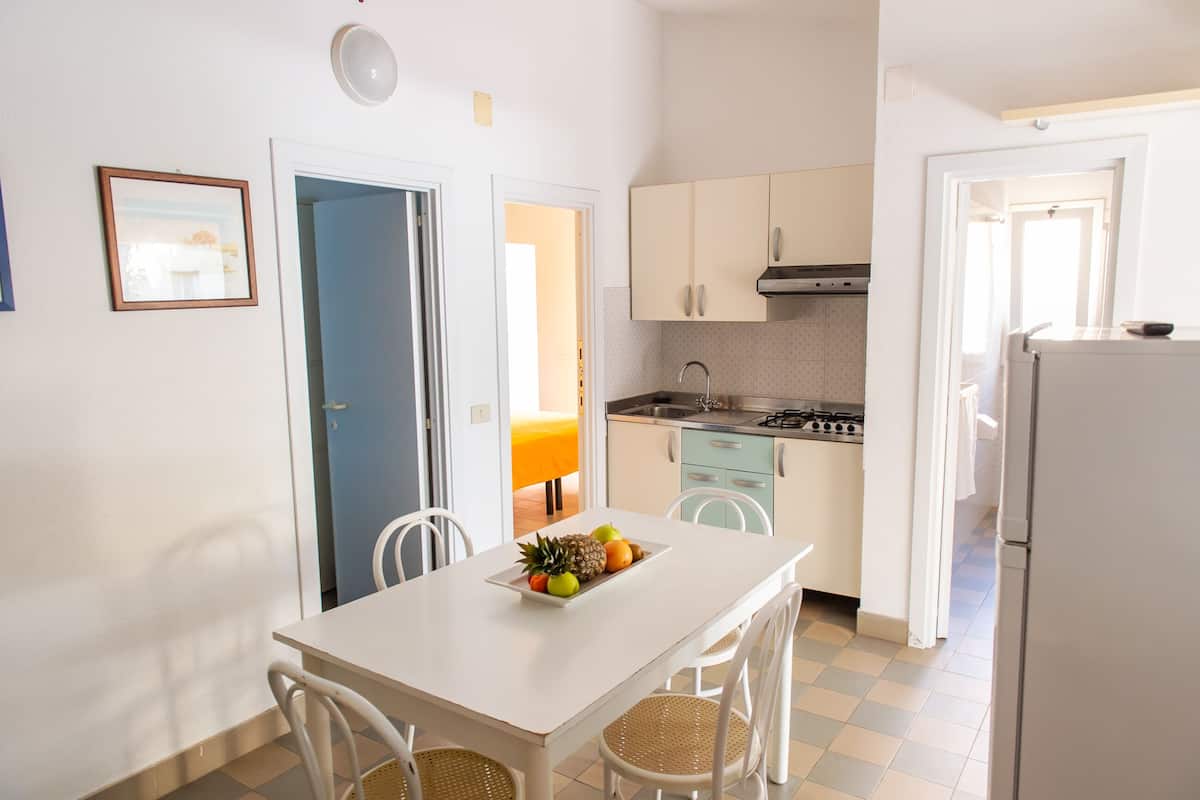 A compact kitchen with white cabinets, a dining table with a fruit bowl, and a refrigerator.