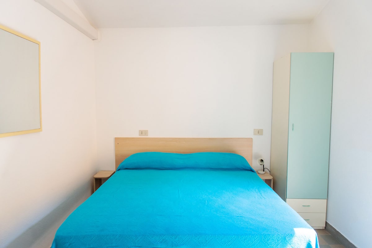 A single bed with a wooden headboard, a teal bedspread, and a nightstand with a lamp. A tall, light green wardrobe is against the wall.