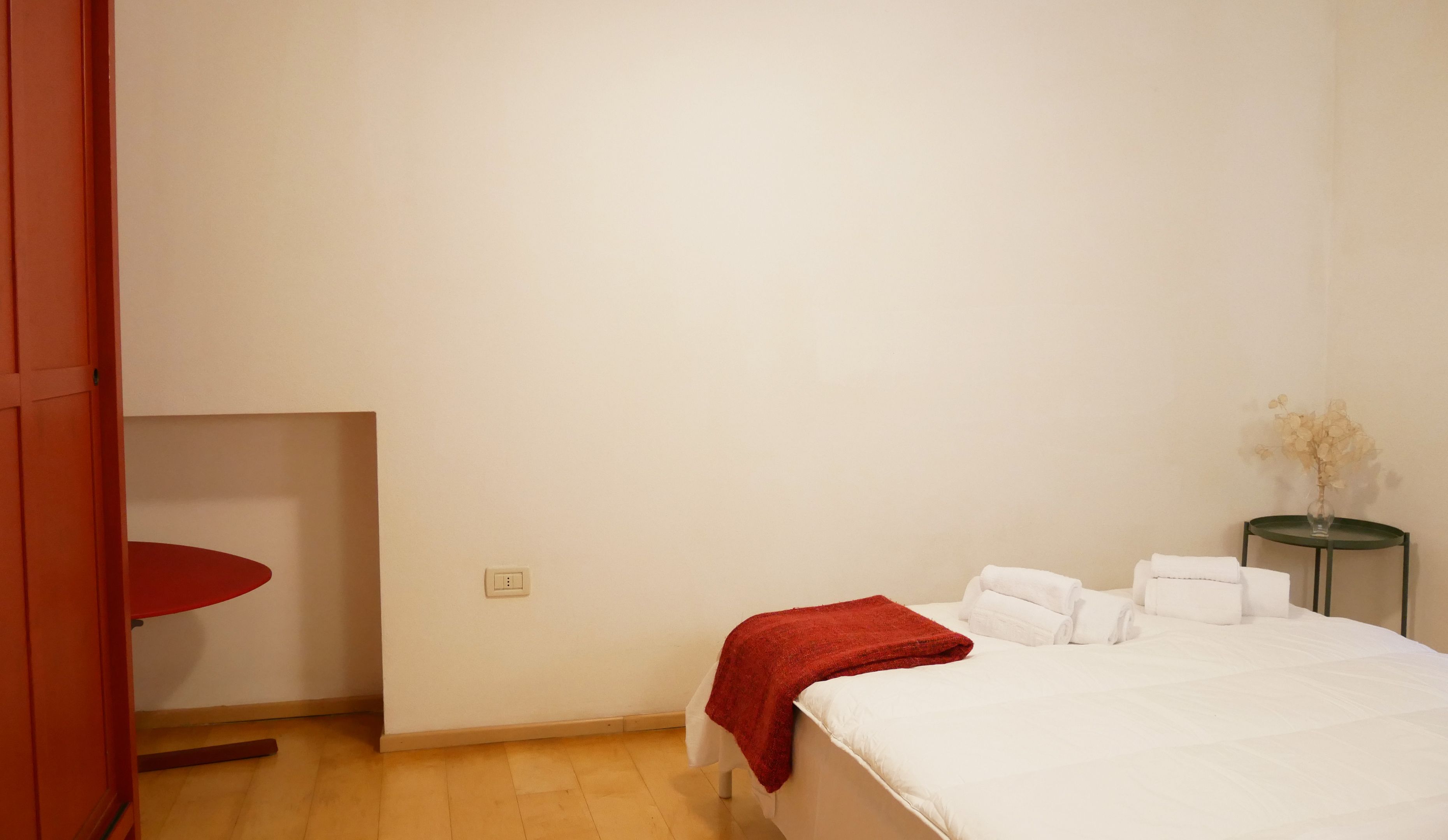 Une chambre d’hôtel avec un lit, une chaise rouge, une petite table avec un vase et un mur blanc.