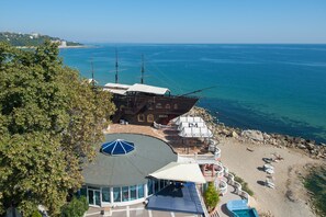A coastal building with a large deck, a circular viewing area, and a beach with sun loungers. - Sirius Beach Hotel & SPA (Varna)