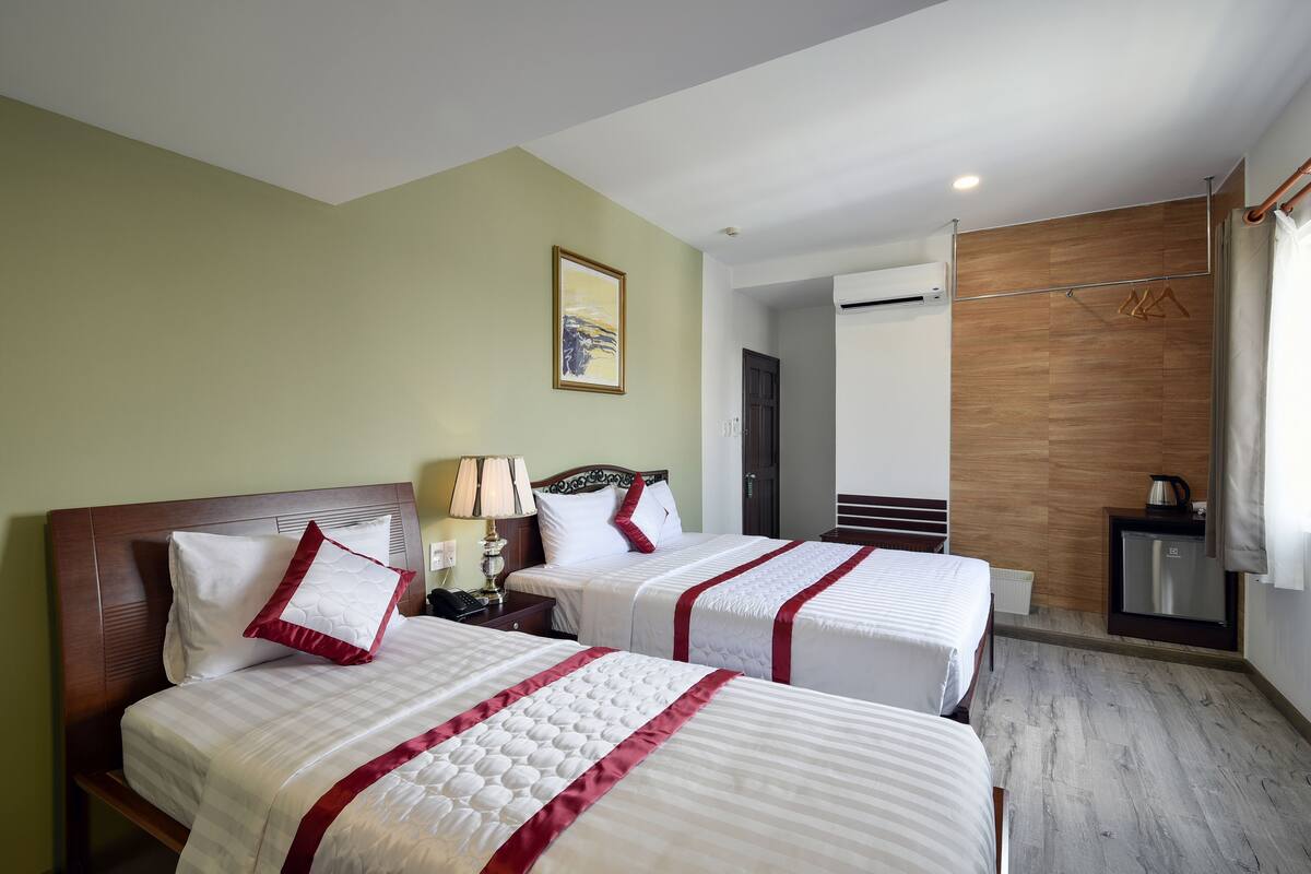 Hotelkamer met twee bedden, een houten hoofdbord, een nachtkastje, een lamp, een airconditioning en een kleine koelkast, plus een houten wandpaneel.