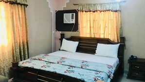 Une chambre à coucher avec un lit en bois, des oreillers blancs, une couvre-lit à motifs floraux, une table de chevet et un climatiseur fixé au mur.