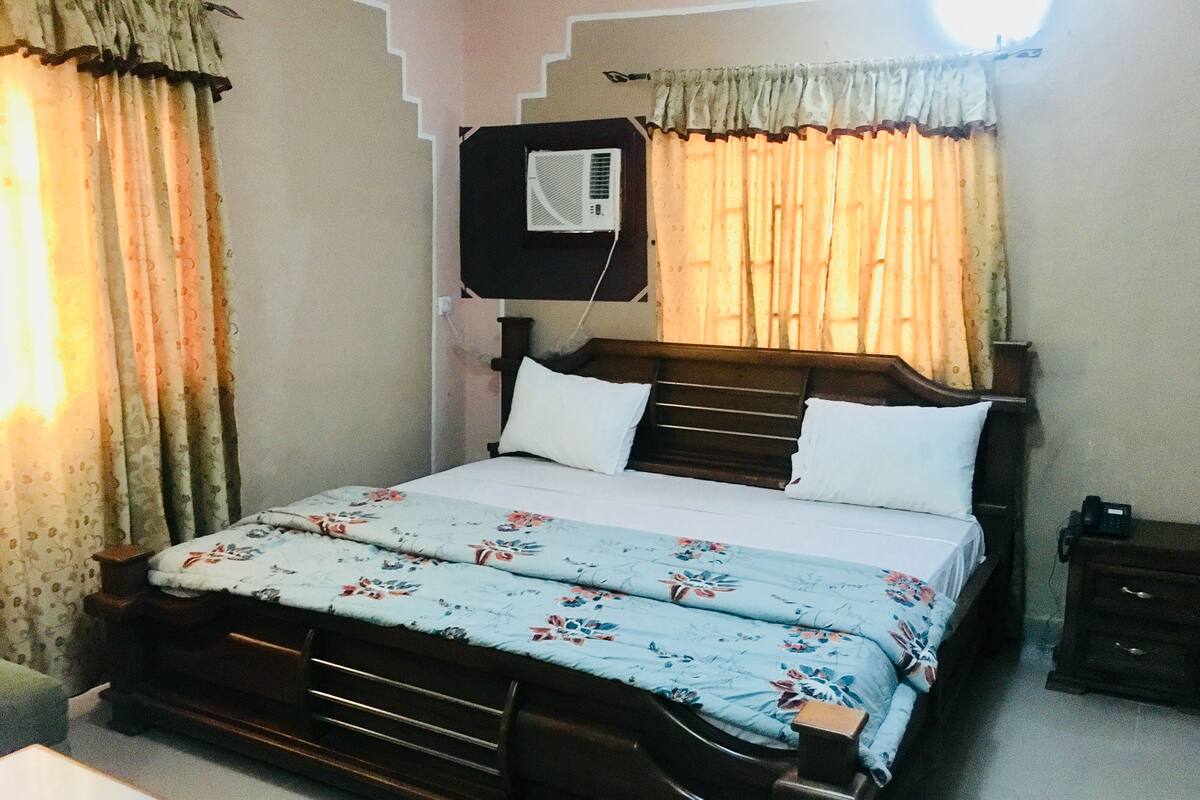 A bedroom with a wooden bed, white pillows, a floral bedspread, a bedside table, and a wall-mounted air conditioner.