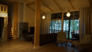 Ein gemütliches Blockhaus-Interieur mit Kamin, Holzwänden, einer Couch, zwei abgedeckten Sesseln und einem großen Fenster mit Blick auf Bäume.