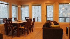 A wooden interior with a dining table and chairs, a sofa, and large windows with a view of trees.