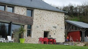 Un edificio de piedra con un gran ventanal y una puerta, mesas al aire libre con sillas rojas y una jardinera verde.