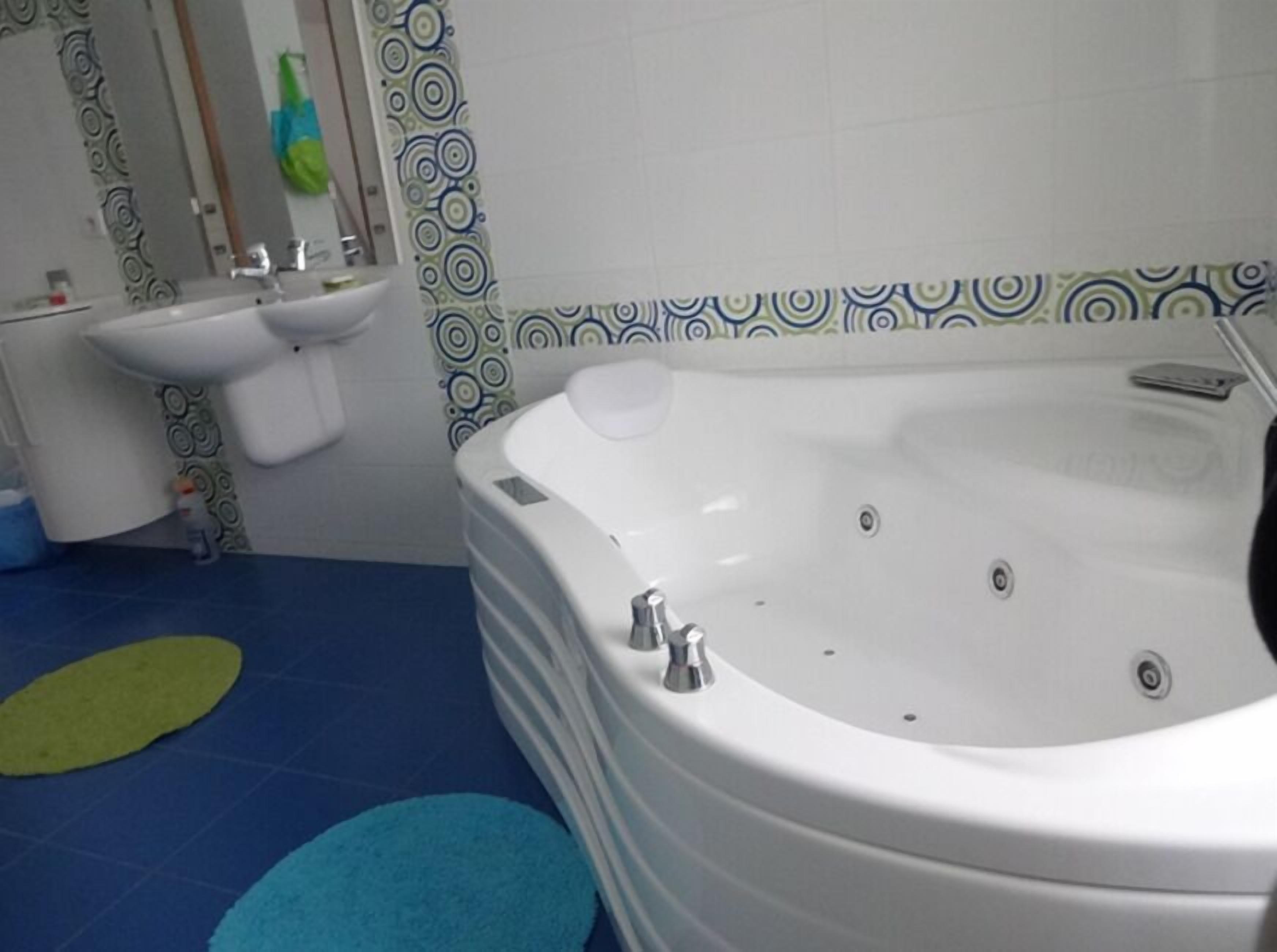 Un baño con una bañera blanca, un lavamanos y un espejo. Las paredes tienen azulejos con diseños y el suelo es azul.