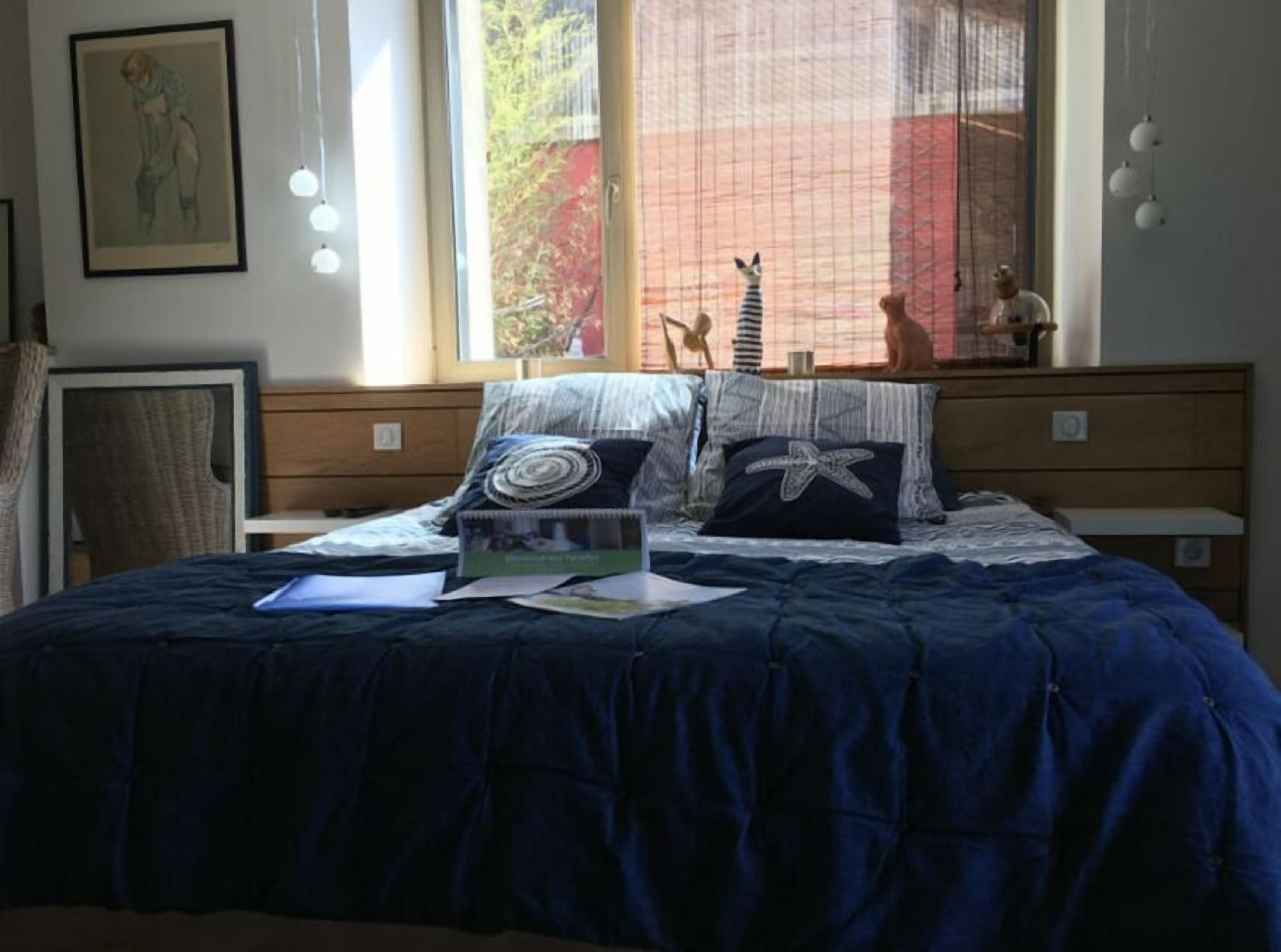 Une chambre avec un grand lit, une fenêtre donnant sur des arbres, un tableau encadré au mur et une petite étagère avec des objets décoratifs.