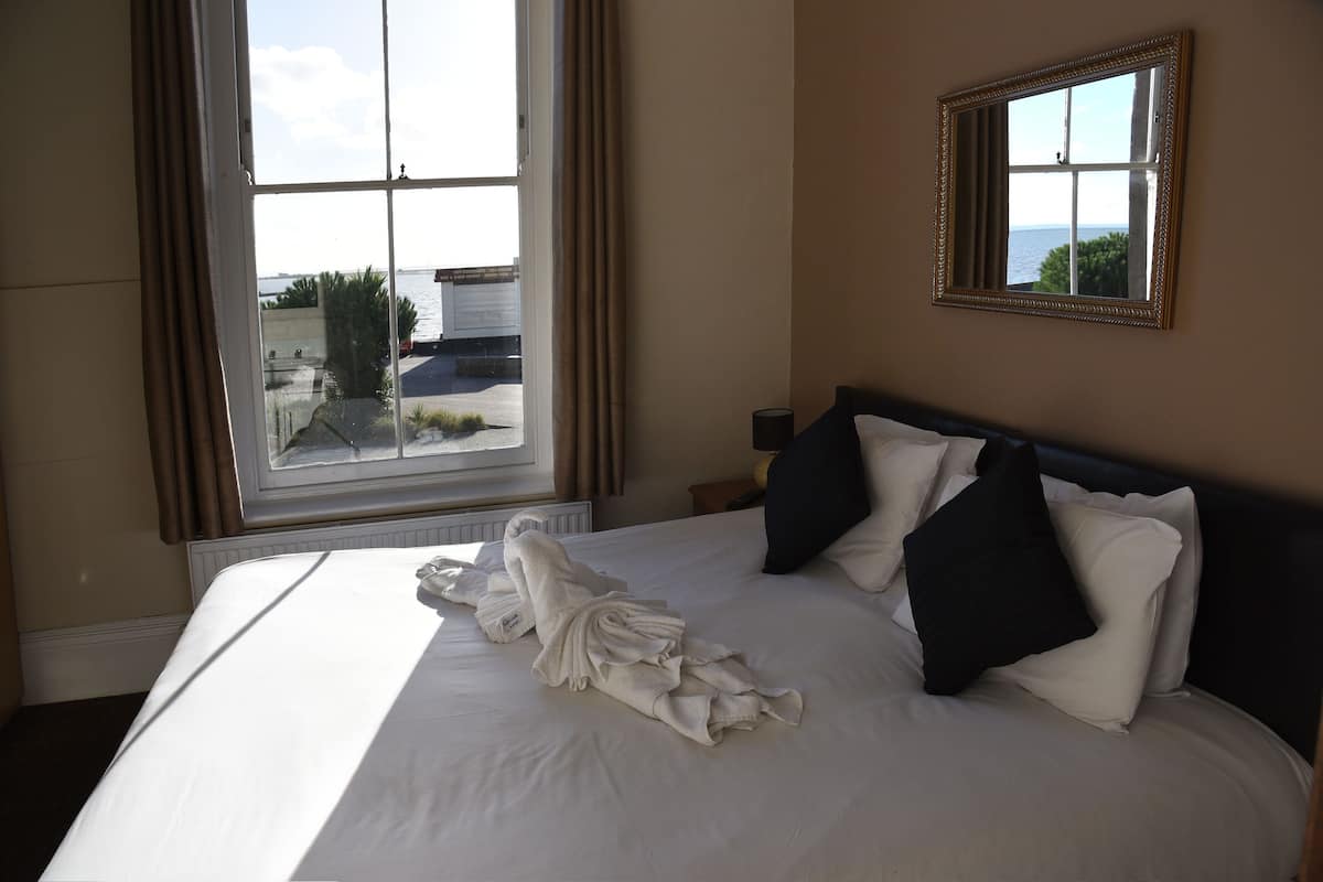 A hotel room with a large bed, a window with curtains, a framed mirror, and a view of the sea.