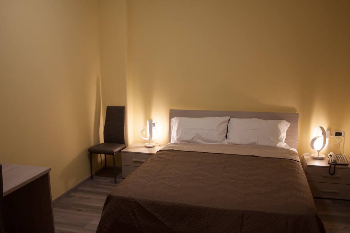 Quarto de hotel com uma cama grande, duas mesas de cabeceira, uma cadeira e uma escrivaninha.