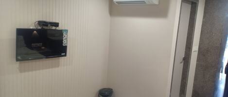 Habitación de hotel con televisor de pantalla plana montado en la pared, equipo de aire acondicionado, basurero y puerta que da a otra habitación.