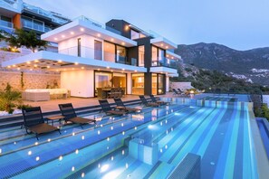 A modern multi-story house with a large outdoor swimming pool, lounge chairs, and a mountain backdrop. - Villa Vogue (Kas)