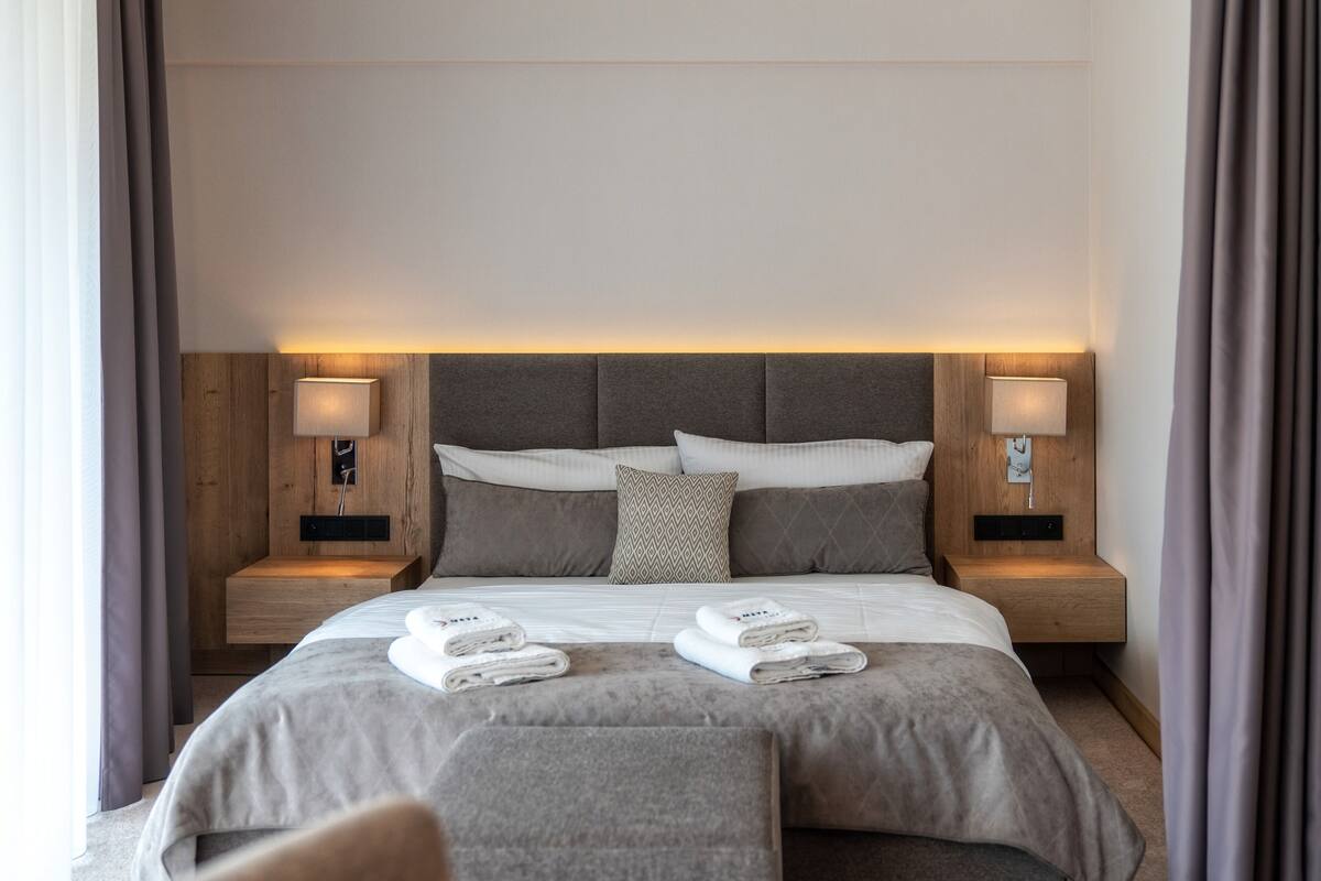 A modern hotel room with a large bed, two bedside tables with lamps, a headboard, and a view of the outside.