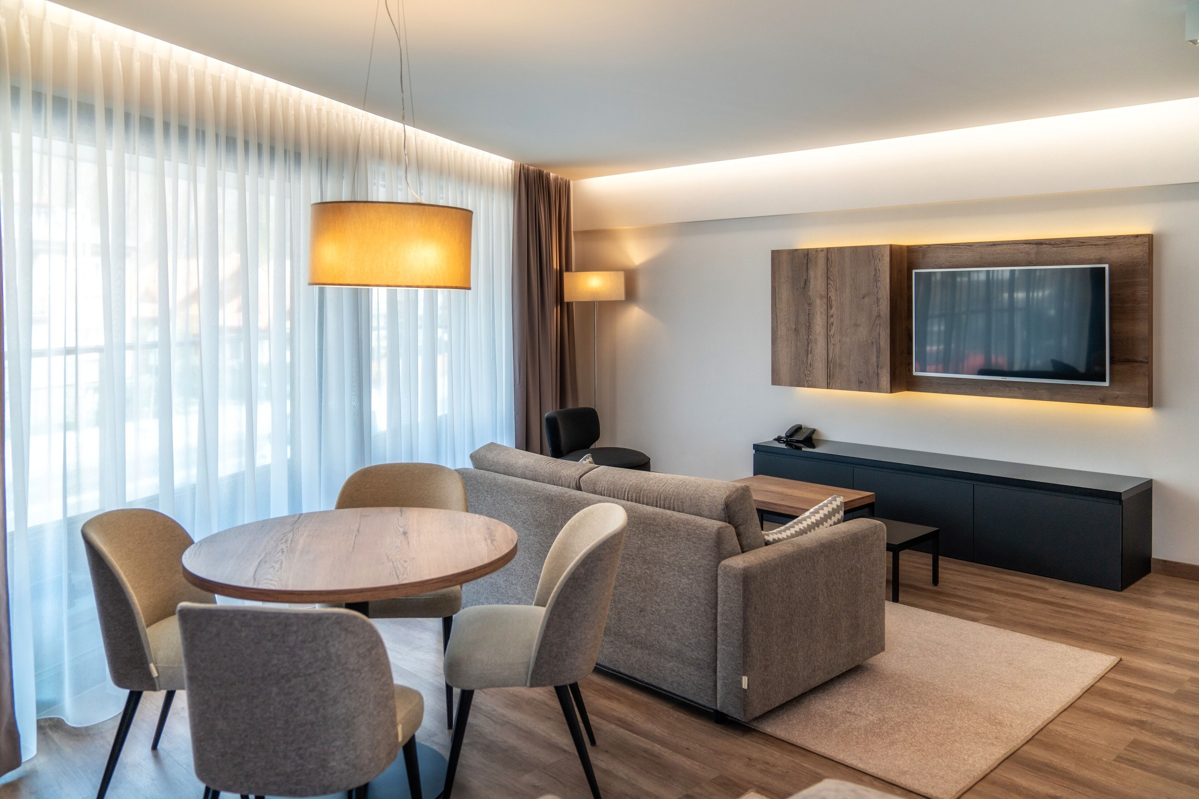 A modern living room with a round wooden table, grey sofa, and a flat-screen TV mounted on the wall.