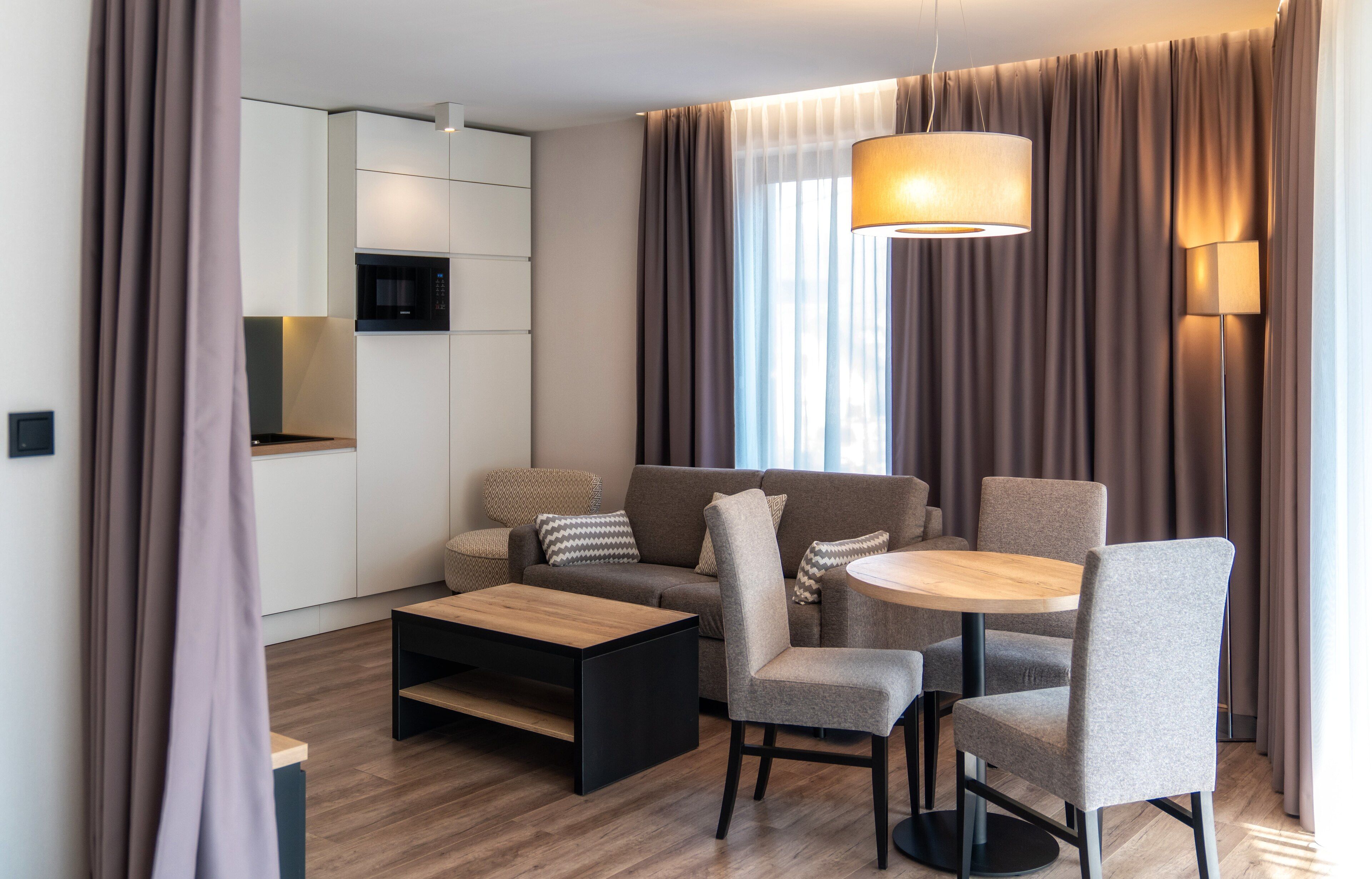 Un salon moderne comprenant un canapé, des fauteuils, une table basse et une table à manger. Il y a également un coin cuisine avec un four à micro-ondes et des placards. La pièce est dotée de grands rideaux et d’un luminaire suspendu.