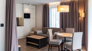 Un salon moderne comprenant un canapé, des fauteuils, une table basse et une table à manger. Il y a également un coin cuisine avec un four à micro-ondes et des placards. La pièce est dotée de grands rideaux et d’un luminaire suspendu.