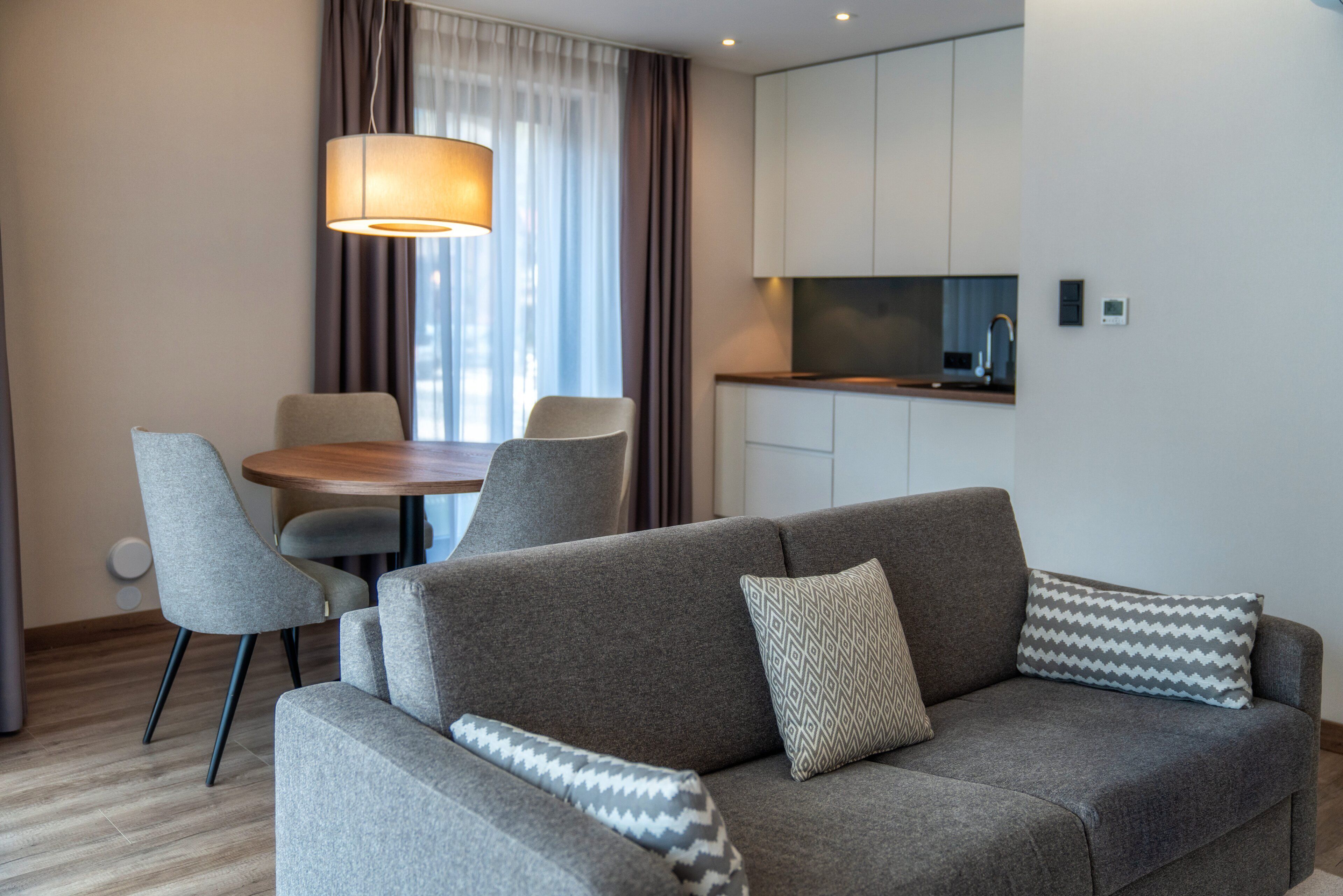 Un salon moderne avec un canapé gris, des coussins à motifs, une table à manger avec des chaises et un coin cuisine doté de placards et d’un plan de travail.
