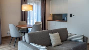 A modern living room with a grey sofa, patterned cushions, a dining table with chairs, and a kitchen area with cabinets and a countertop.