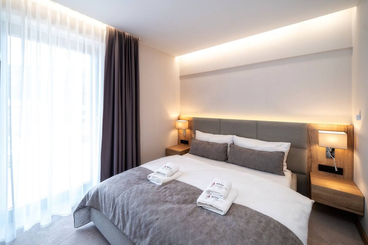 A hotel room with a large bed, two bedside tables with lamps, a headboard, and a window with sheer curtains.