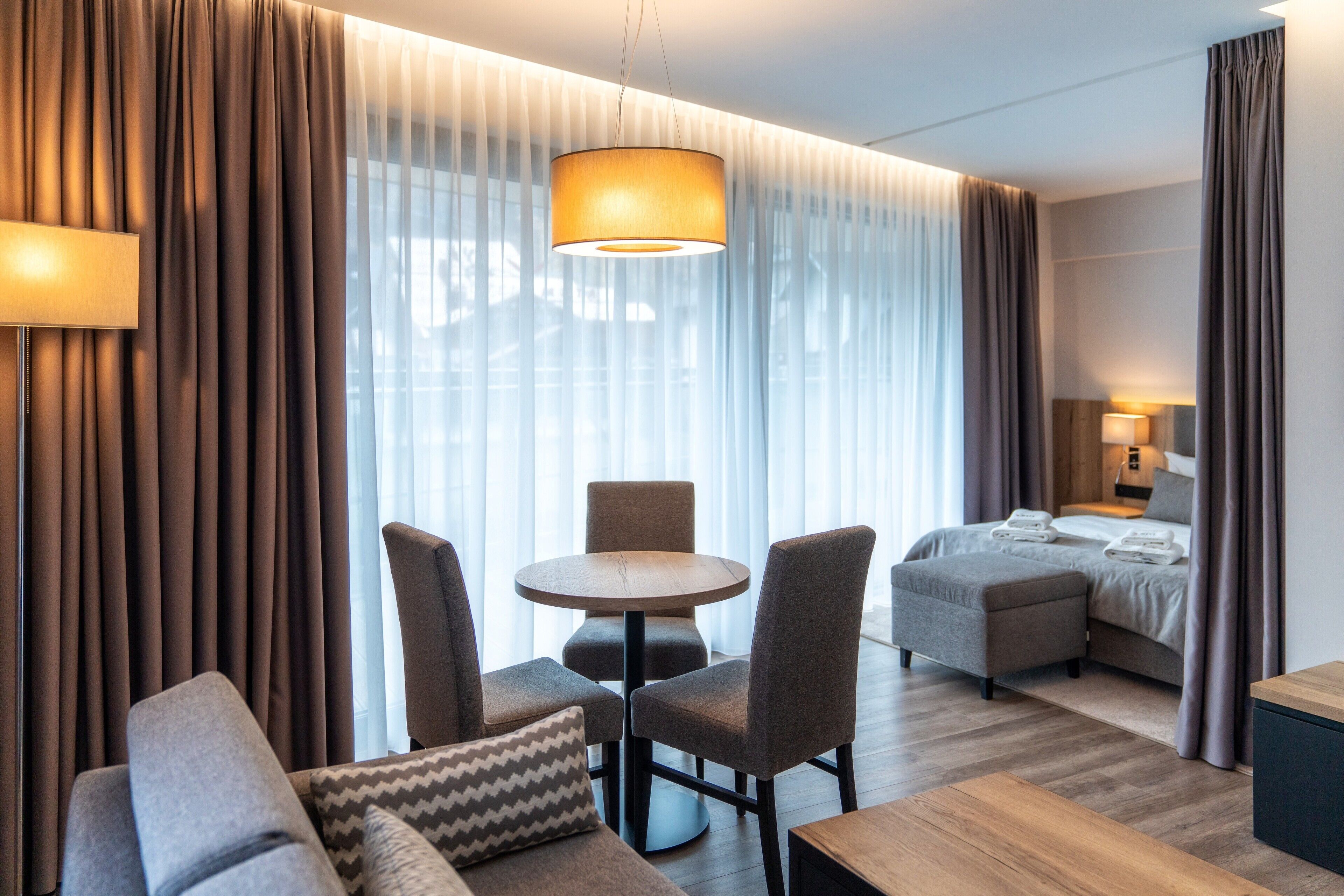 Una habitación de hotel moderna con cama, zona de estar con mesa y sillas, y un ventanal con cortinas.