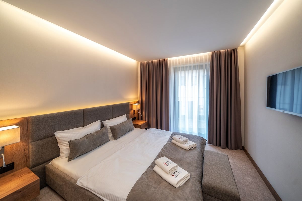 A modern hotel room with a large bed, bedside tables, a flat-screen TV, and a window with curtains.
