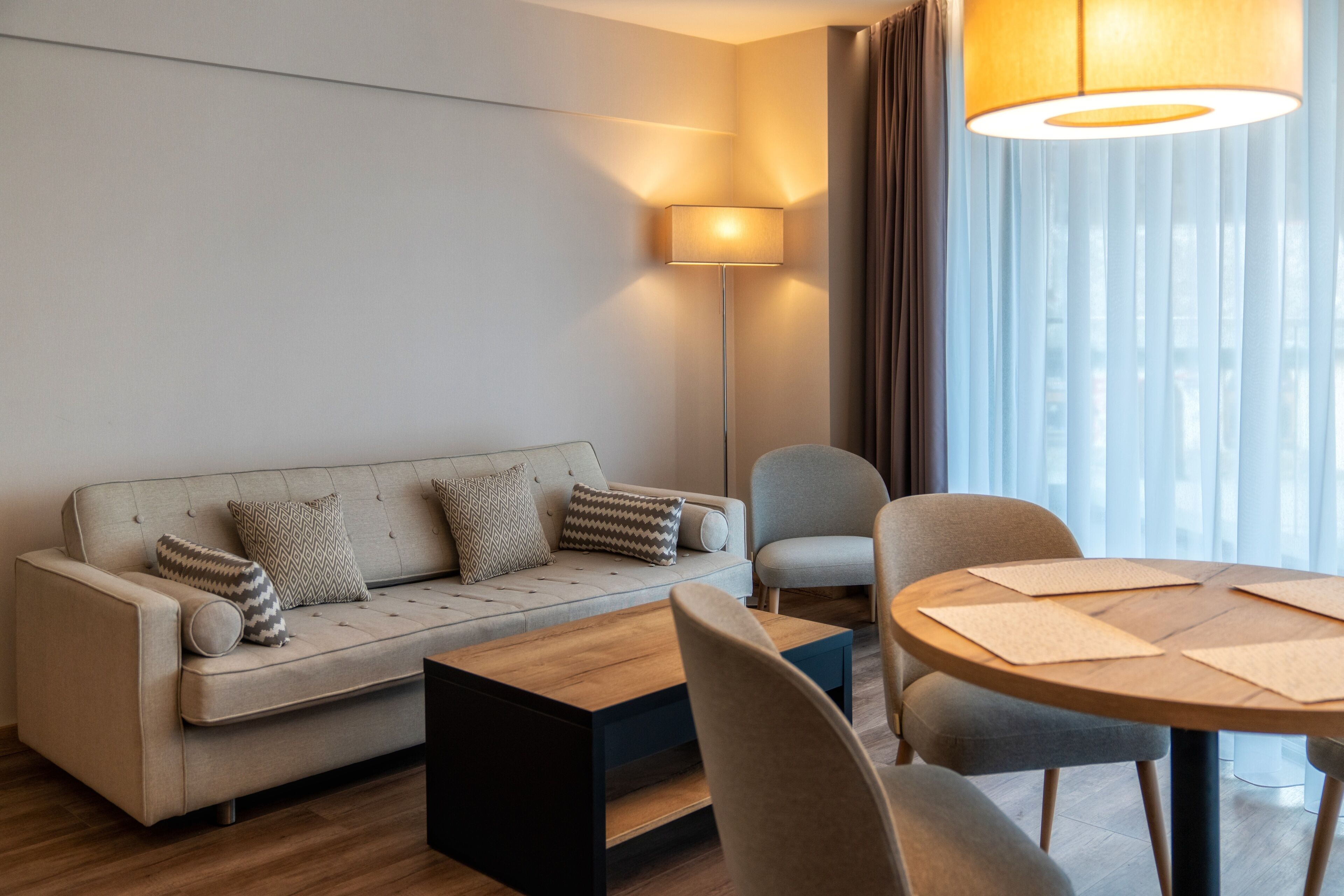A modern hotel room with a sofa, a coffee table, a dining table with chairs, a floor lamp, and a view of the city through sheer curtains.