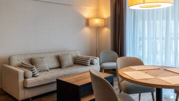A modern hotel room with a sofa, a coffee table, a dining table with chairs, a floor lamp, and a view of the city through sheer curtains.